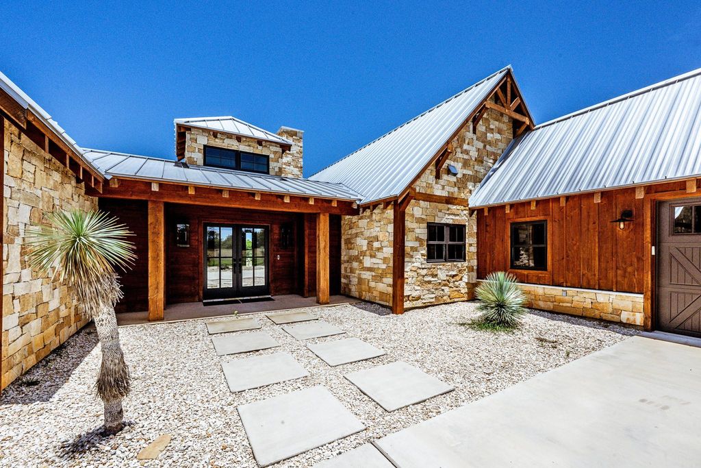 Courtyard entrance of a Texas Hill Country-style home with stone and wood siding, metal roof, and stepping stone path through gravel landscaping