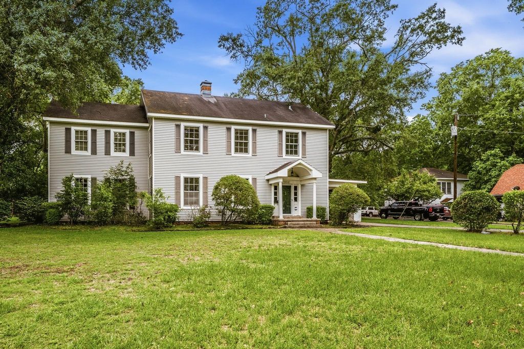 Two-story white colonial-style home with shuttered windows, covered entry, chimney, and large green lawn surrounded by mature trees