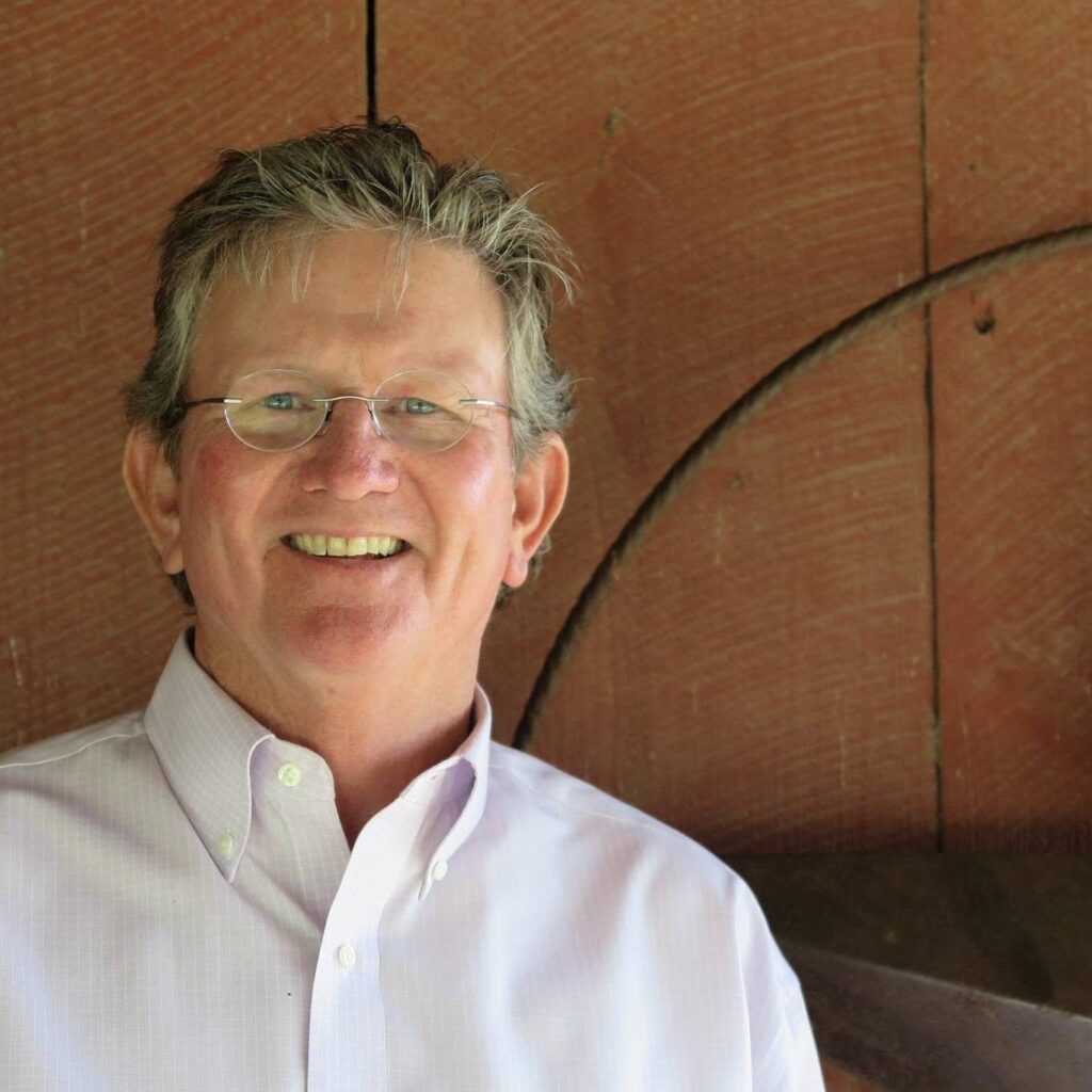 Smiling Sonny Tunstall with gray hair and glasses wearing a light pink button-down shirt, posed in front of a wooden wall