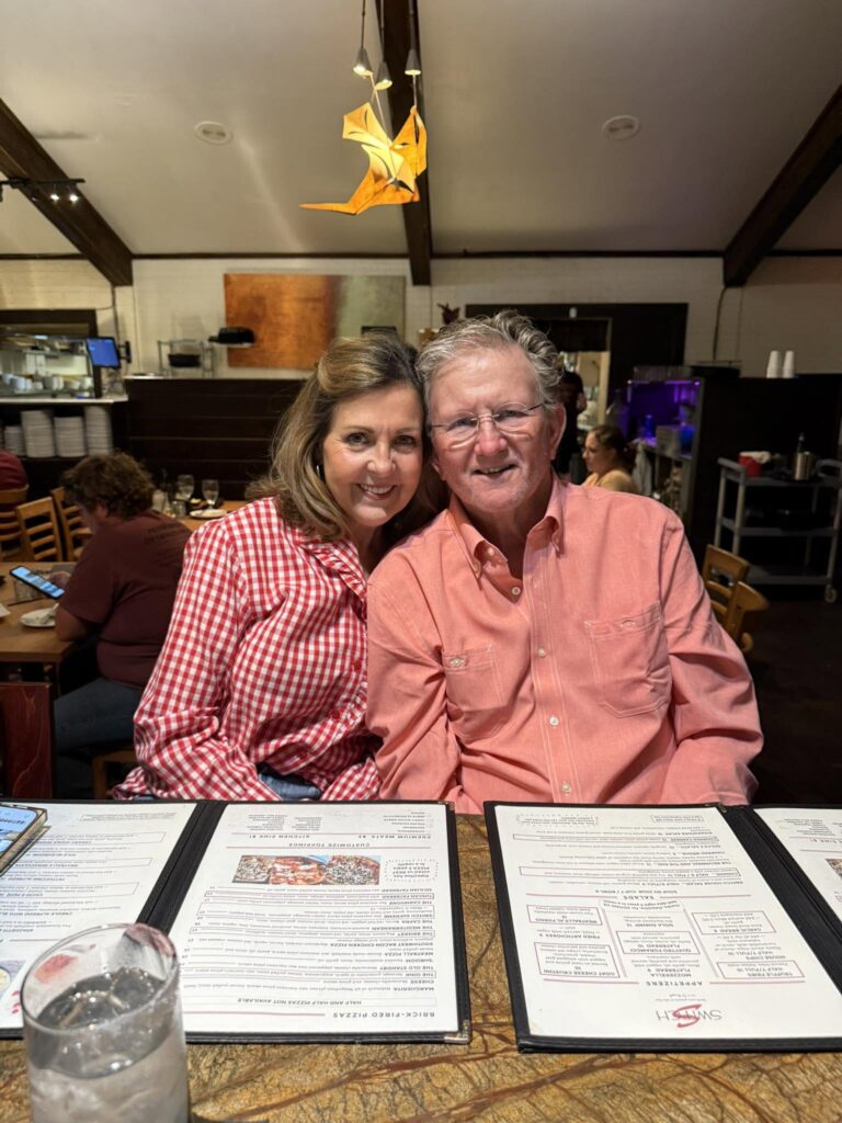 A woman in a red gingham shirt and an older man in a salmon shirt smile together at a restaurant table with menus in front of them