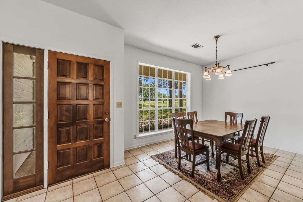Dining area with wood table and chairs on a patterned rug, beside a large window, with a wood panel front door and sidelights nearby