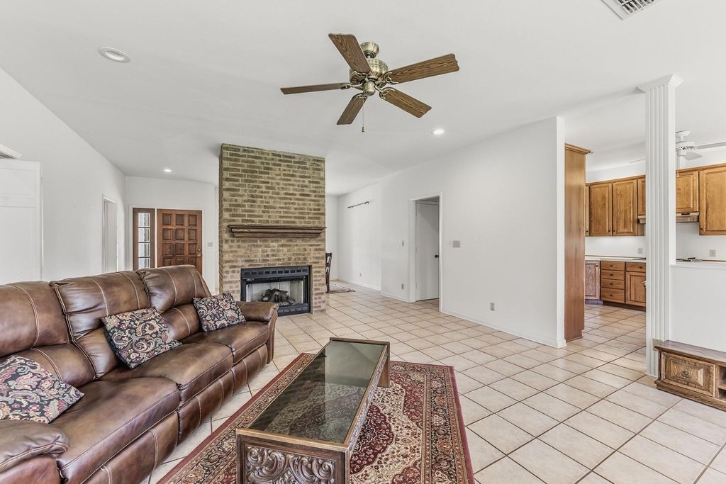 Spacious living room with leather sofa, brick fireplace, ceiling fan, tile floors, and open view to kitchen