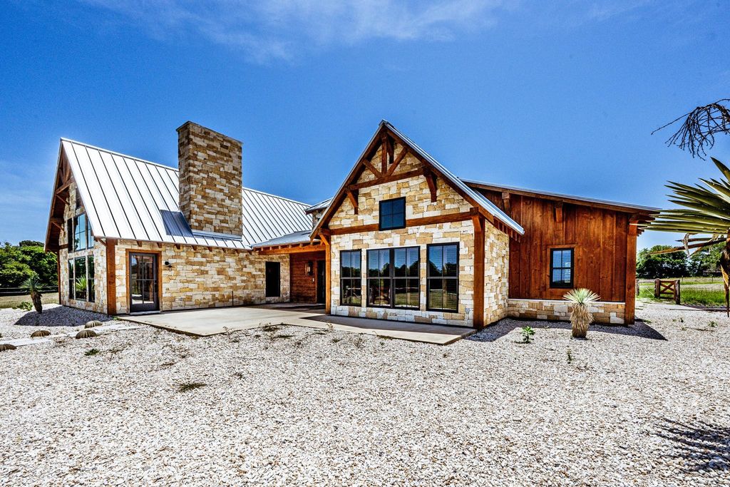 Exterior of a Texas Hill Country-style home with stone and wood siding, metal roof, stone chimney, and gravel yard